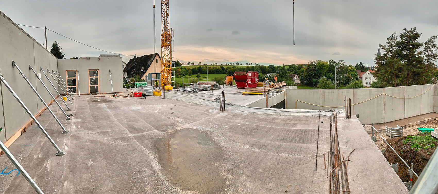 Baustelle auf einer Betonplatte mit Kran, Bauholz, Gerüsten; Arbeiter und ländliche Umgebung.