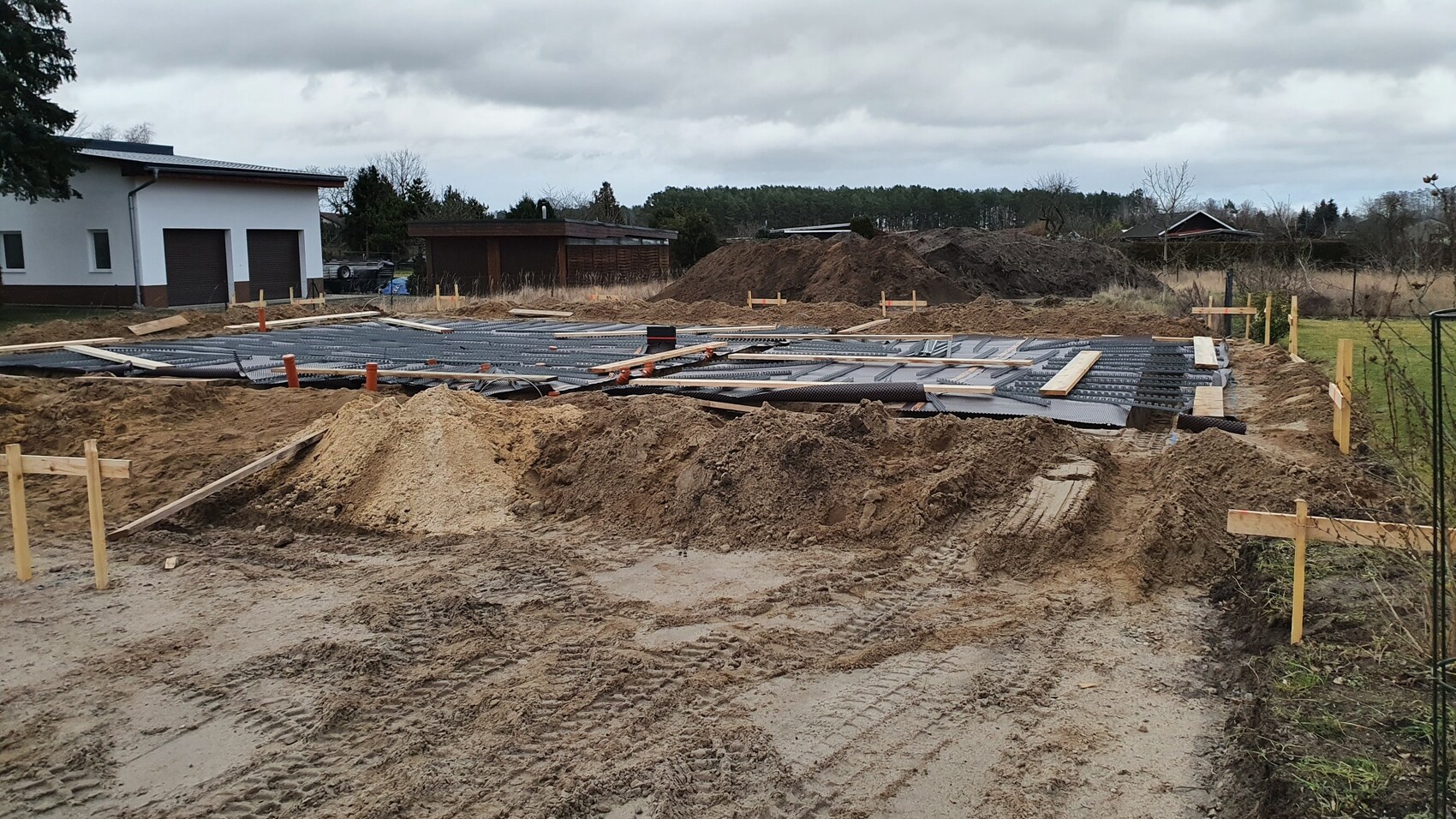 Baustelle: Fundamentplatte wird vorbereitet, Erdhaufen, Holzpfosten, Haus im Hintergrund.