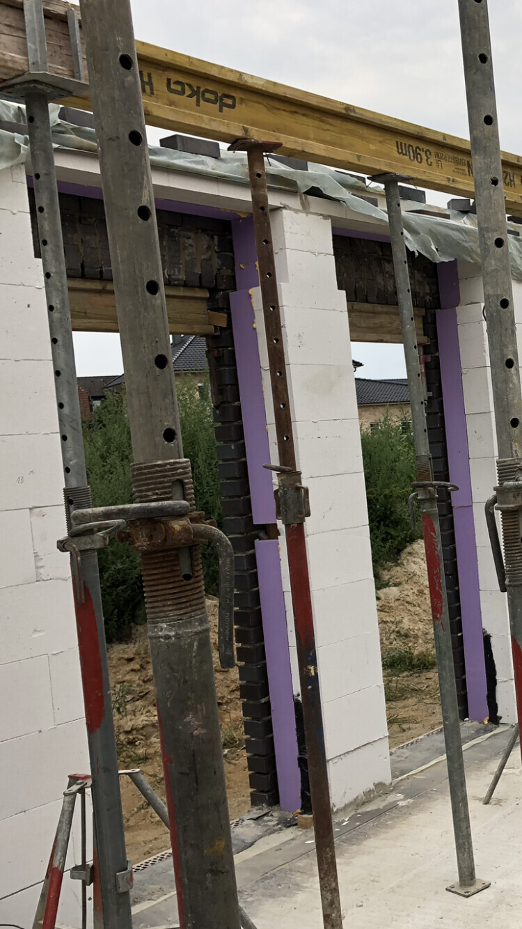 Baustelle mit Metallstützen, Holzbalken und weißen Mauerblöcken; violette Dämmplatten sichtbar.