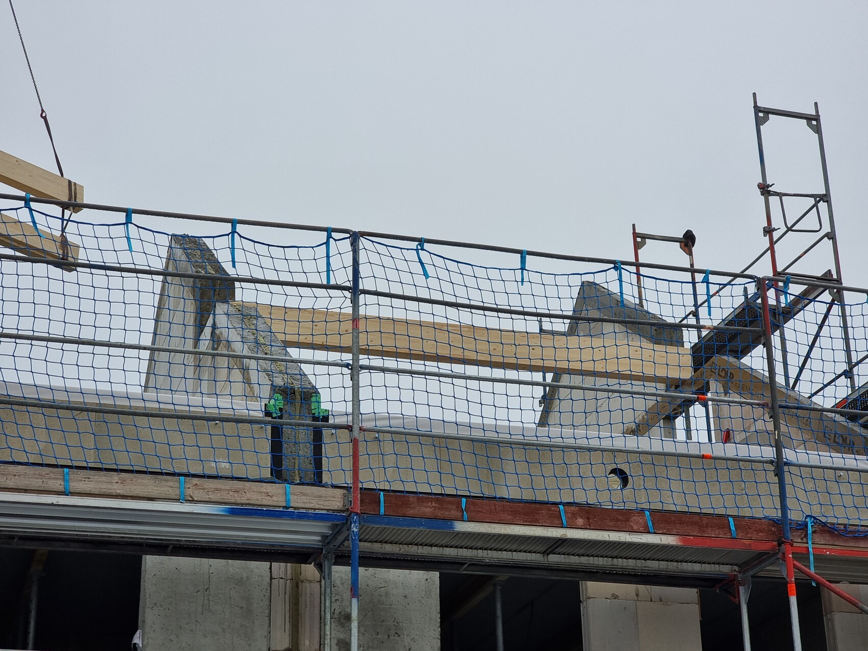 Baustellenszene: Gerüst mit blauen Sicherheitsnetzen, Holzbalken und Betonbauteil vor grauem Himmel