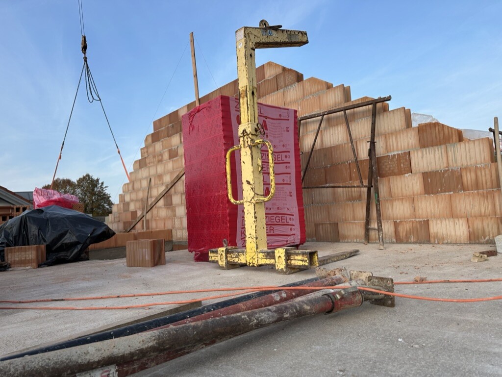 Baustelle mit gestapelten Ziegeln, gelber Hebevorrichtung und rosa verpacktem Block vor Mauer