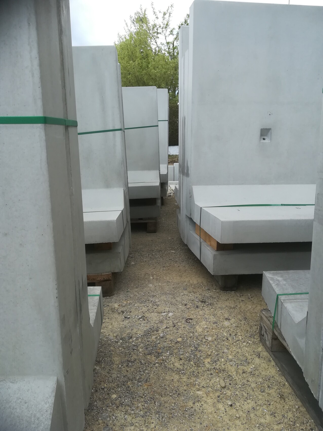 Große graue Betonplatten stapeln sich auf Paletten, grüne Bänder, Durchgang dazwischen.