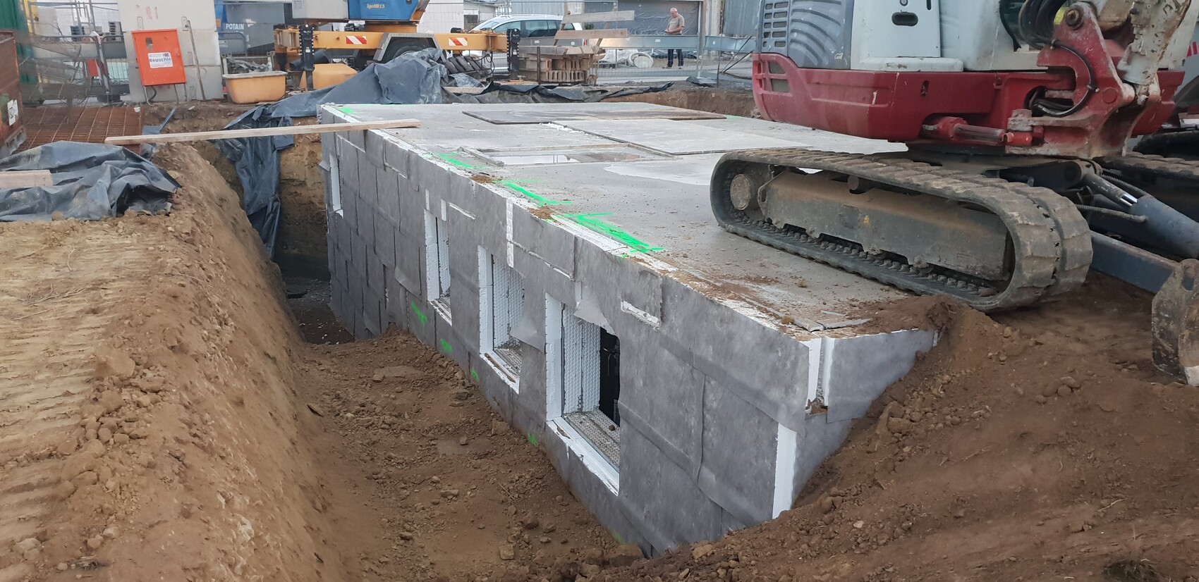 Baustelle mit Erdgraben und Betonblockwand; Bagger mit Raupenfahrwerk rechts.
