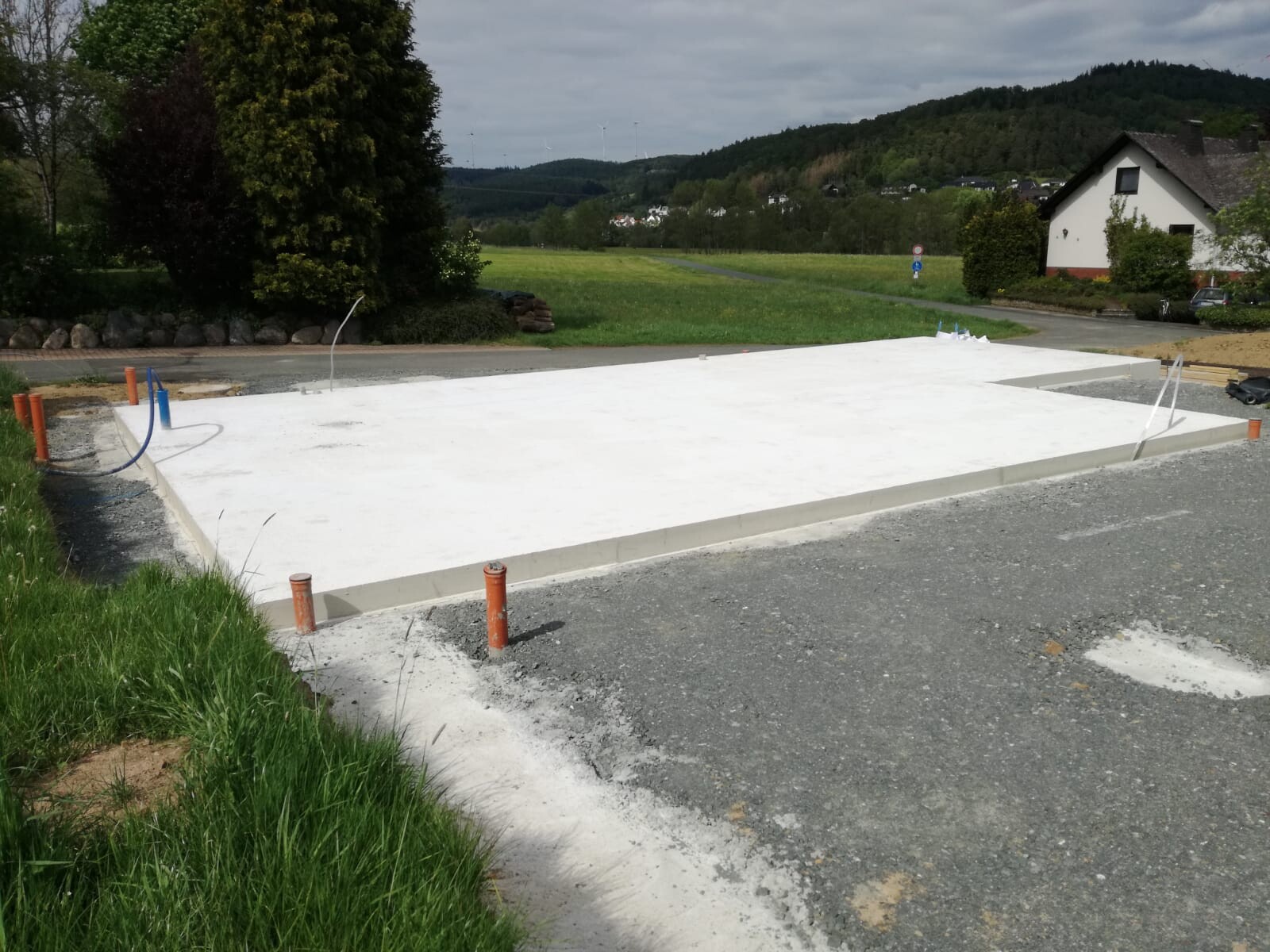 Weiße Beton-Grundplatte auf Baufläche mit Rohren; ländliche Umgebung.