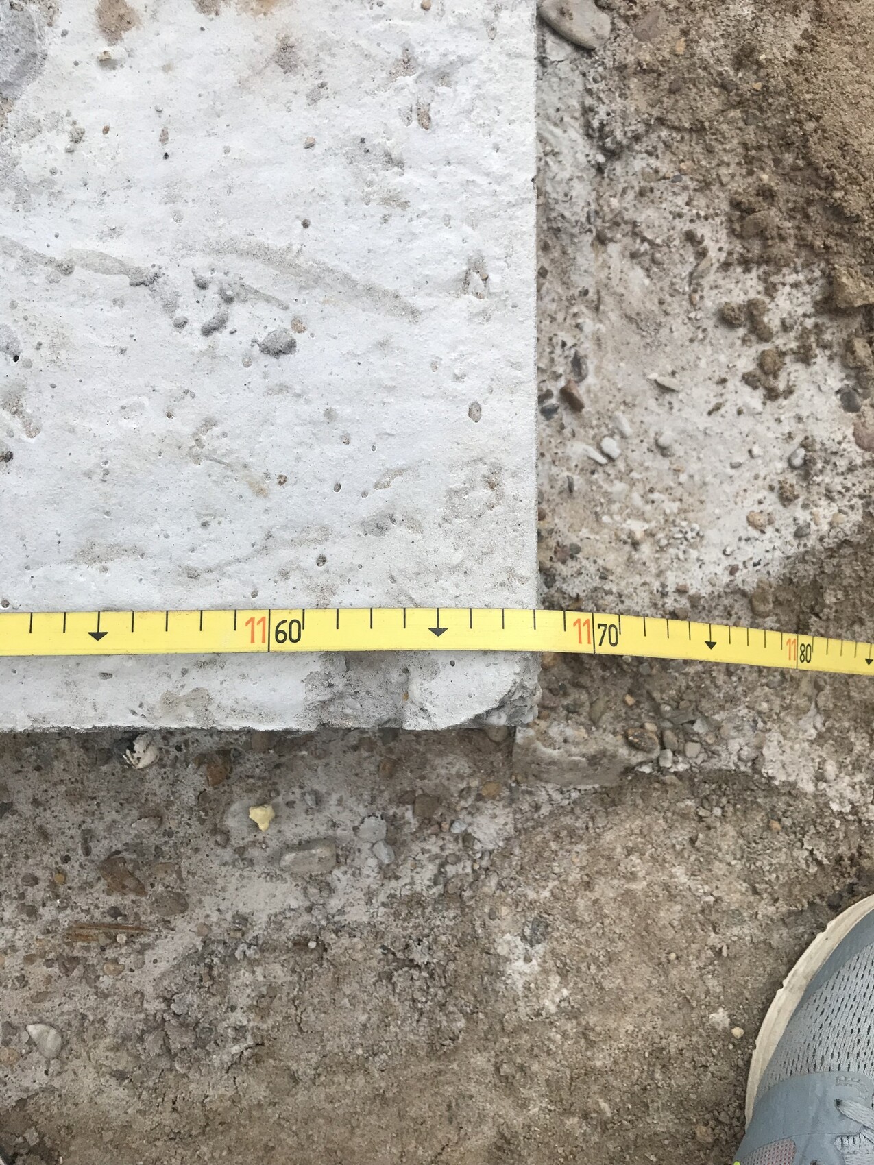Baustelle: Betonplatte liegt auf Erde, Messband zeigt ca. 60–70 cm, Schuh am Rand sichtbar.