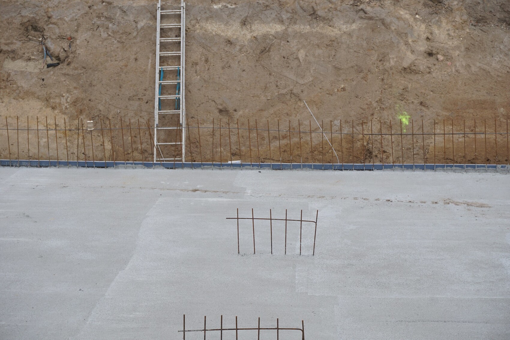 Baustelle: Leiter lehnt an Erdwall, Bewehrung ragt aus Betonboden, grüne Markierung sichtbar.