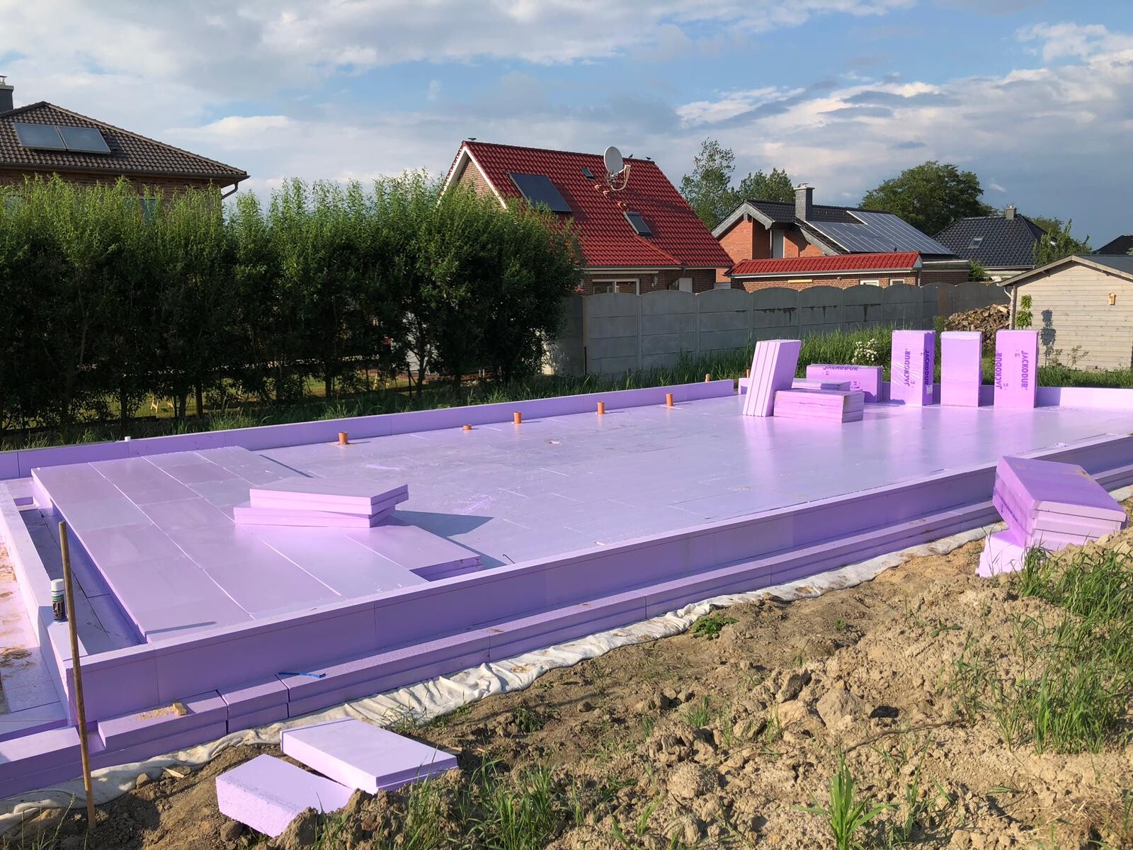 Fundamentbau einer Häuserbaustelle mit violetten Dämmplatten, Nachbarsgärten im Hintergrund