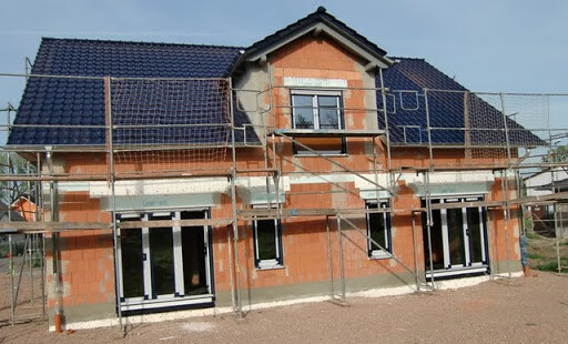 Zweistöckiges Haus im Rohbau mit Backsteinfassade, blauem Dach und Gerüst rundum.