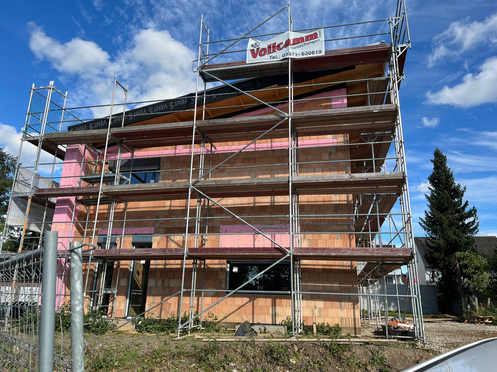 Mehrstöckiges Gebäude im Bau mit Gerüst, Backsteinmauer und pinken Dämmplatten vor blauem Himmel.
