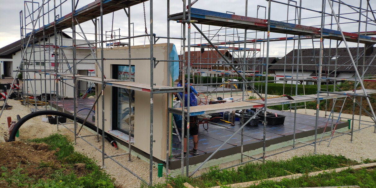 Baustelle mit Gerüst um einen Neubau; Arbeiter arbeiten an der Bodenplatte.