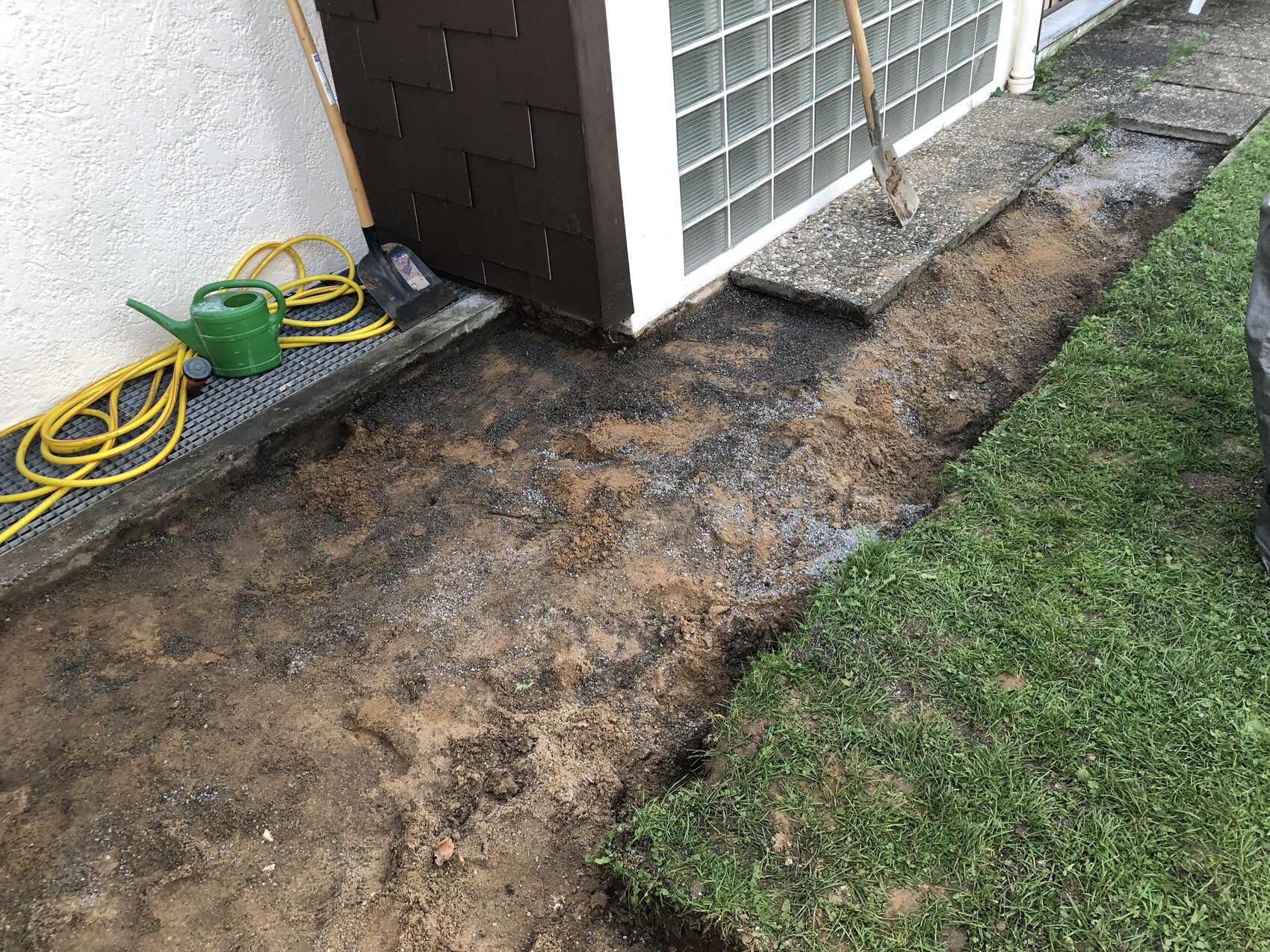 Graben neben Hauswand: ausgehobene Erdgrube mit Sand, Schaufel, Gartenschlauch und Gießkanne.