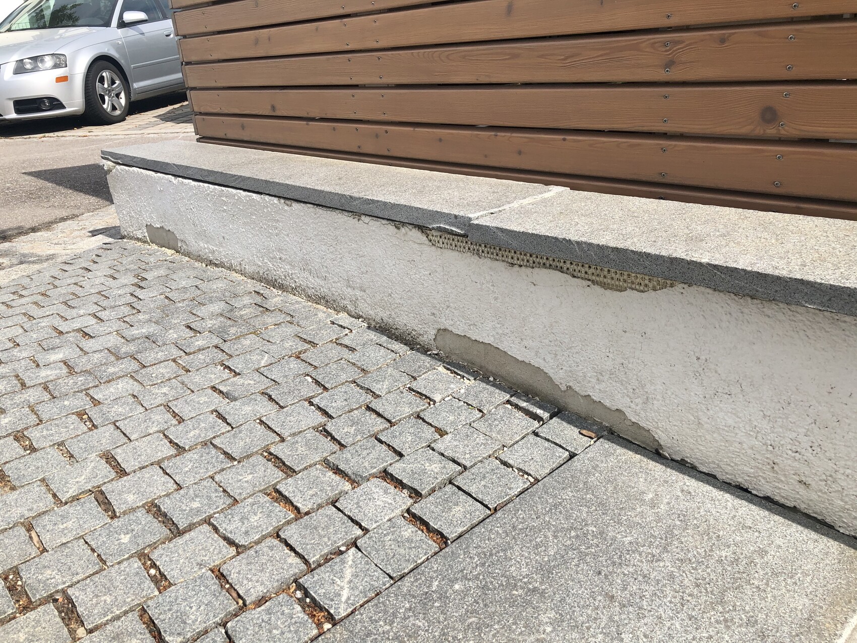 Betonbank vor Holzverkleidung, gepflasterter Vorplatz und silbernes Auto im Hintergrund.