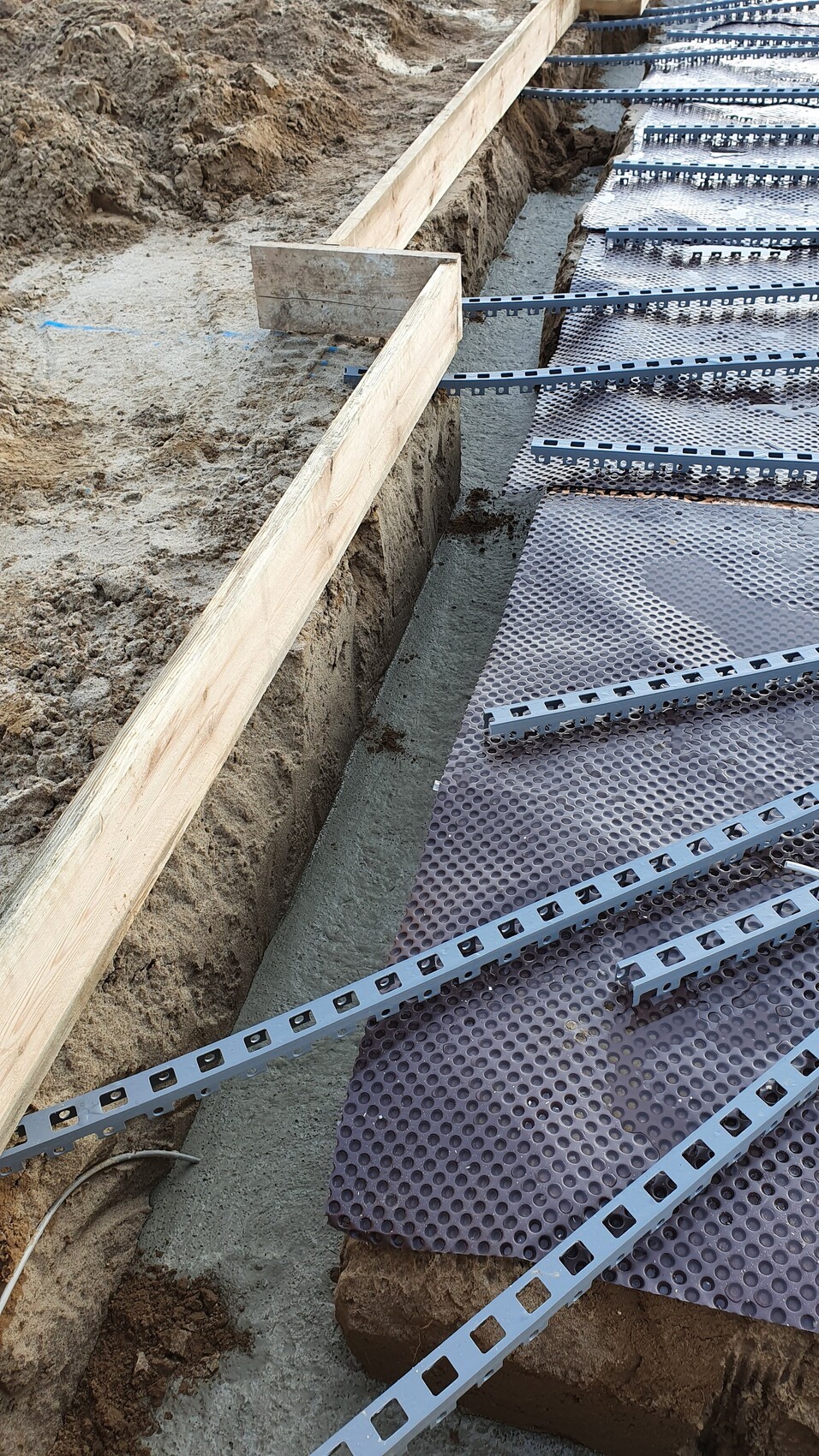 Baugrube mit Holzformrahmen, quer liegende gelochte Metallrinnen und rutschfeste Matte.