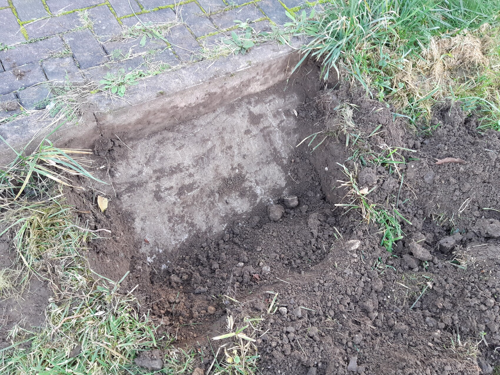 Freigelegter Erd- und Fundamentbereich neben gepflastertem Gehweg mit Gras