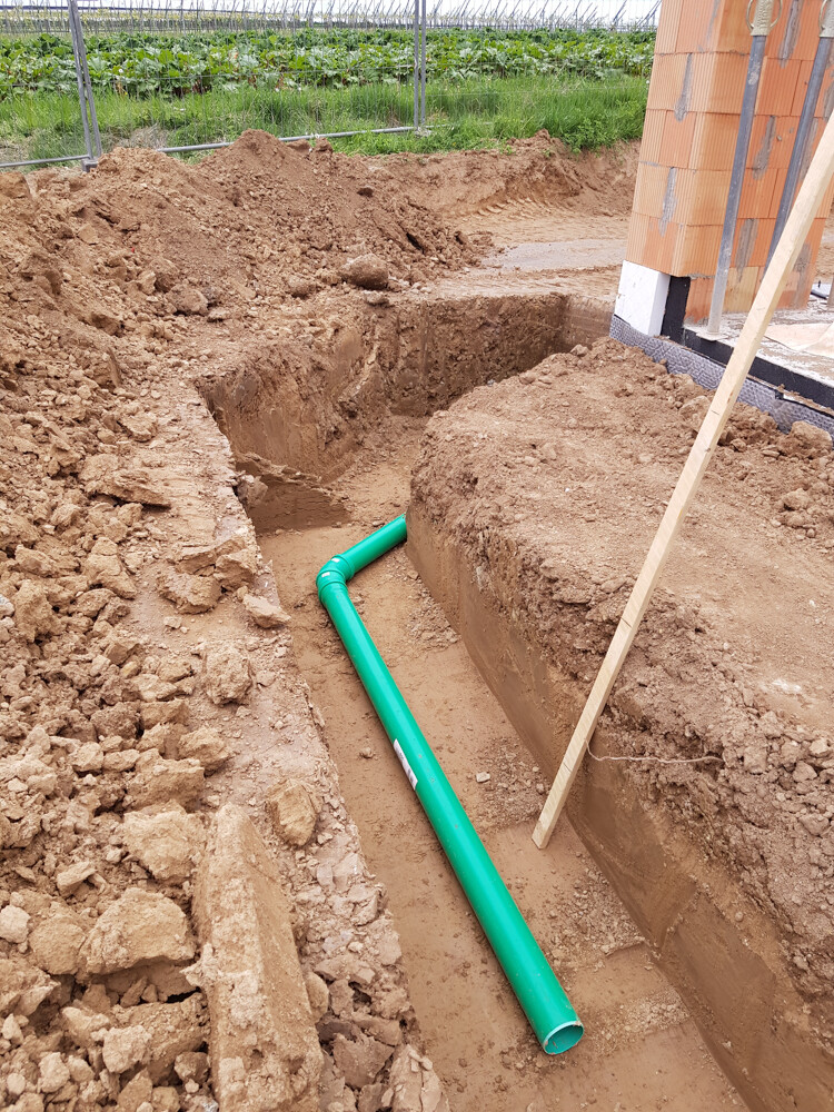 Graben mit grünem PVC-Rohr in einer Baugrube neben dem Hausfundament.