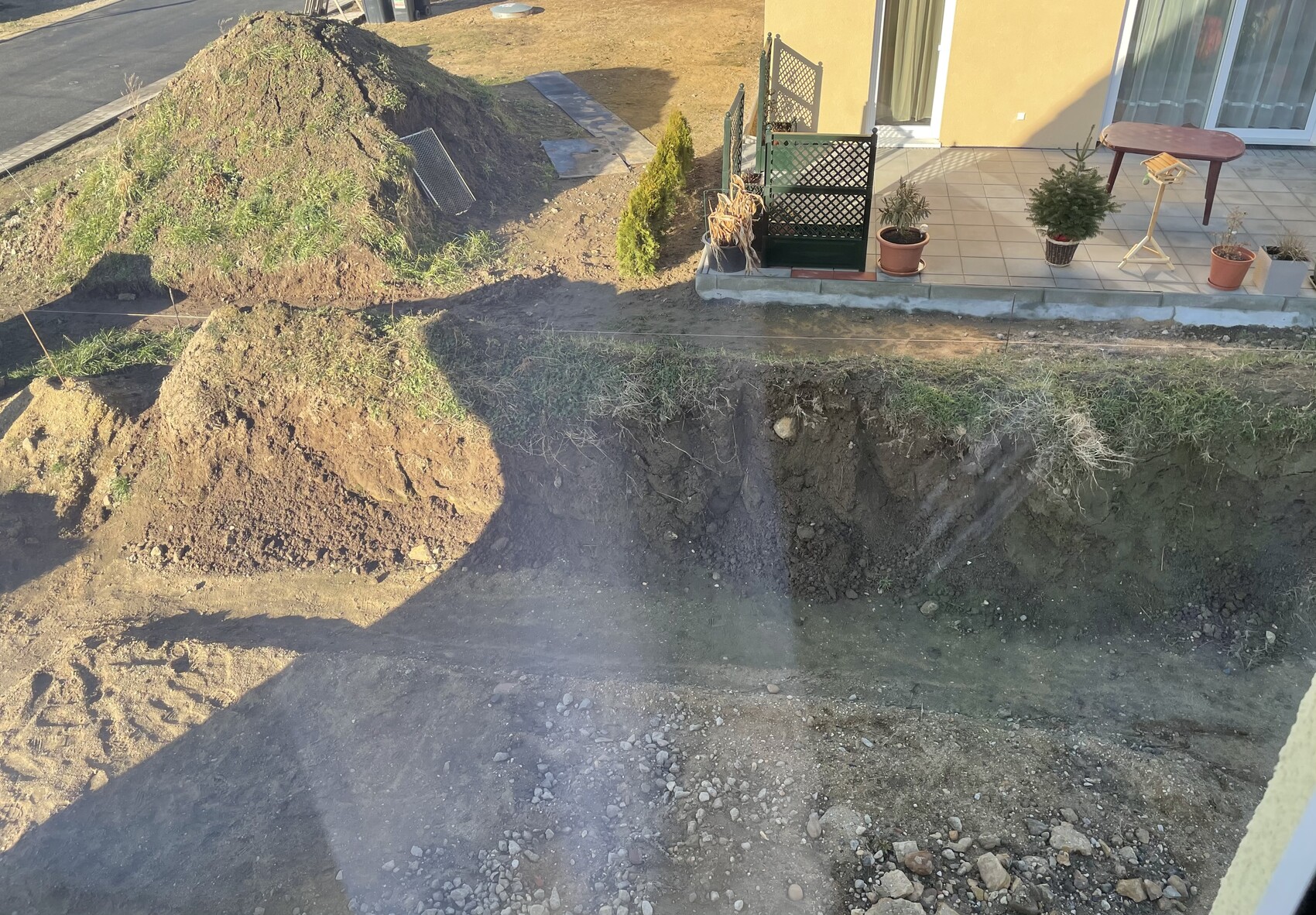 Tief ausgehobene Baugrube am Haus, Erdhügel neben der Terrasse mit Topfpflanzen.