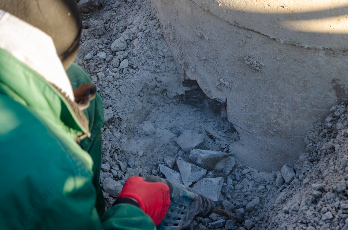 Person in grünem Mantel und roten Handschuhen zerschlägt Gestein mit Bohrhammer in einem Graben.