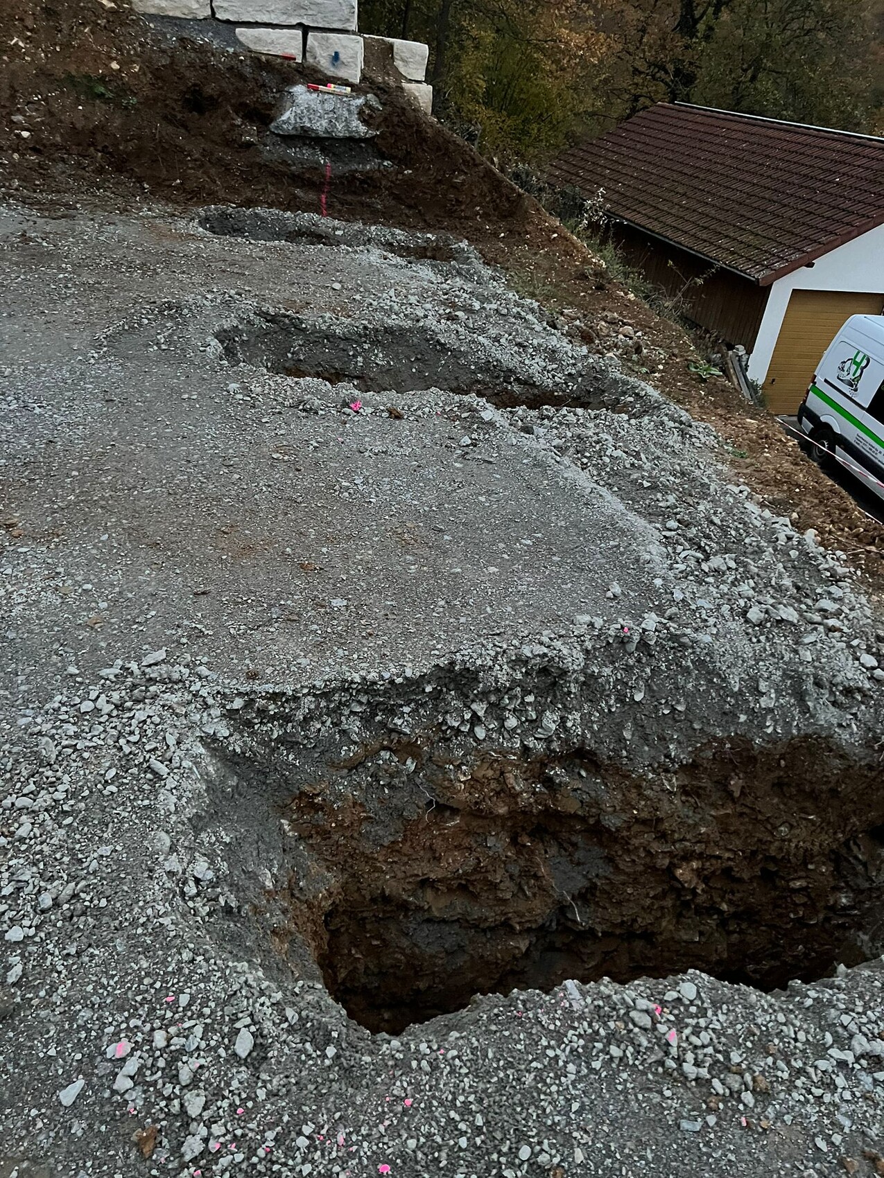 Große Grube auf Baustelle mit Kies; links Betonblöcke, rechts Garage und Lieferwagen.