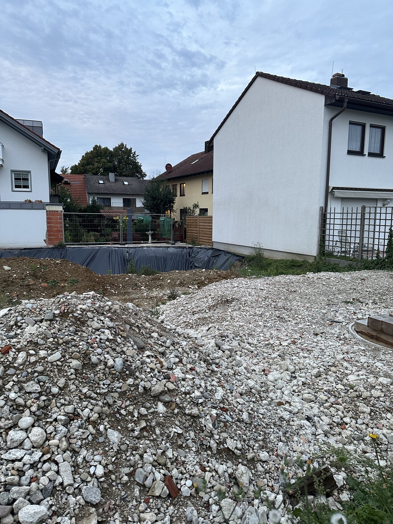 Große Baugrube im Wohngebiet, davor Kies- und Geröllhaufen, angrenzende Wohnhäuser.