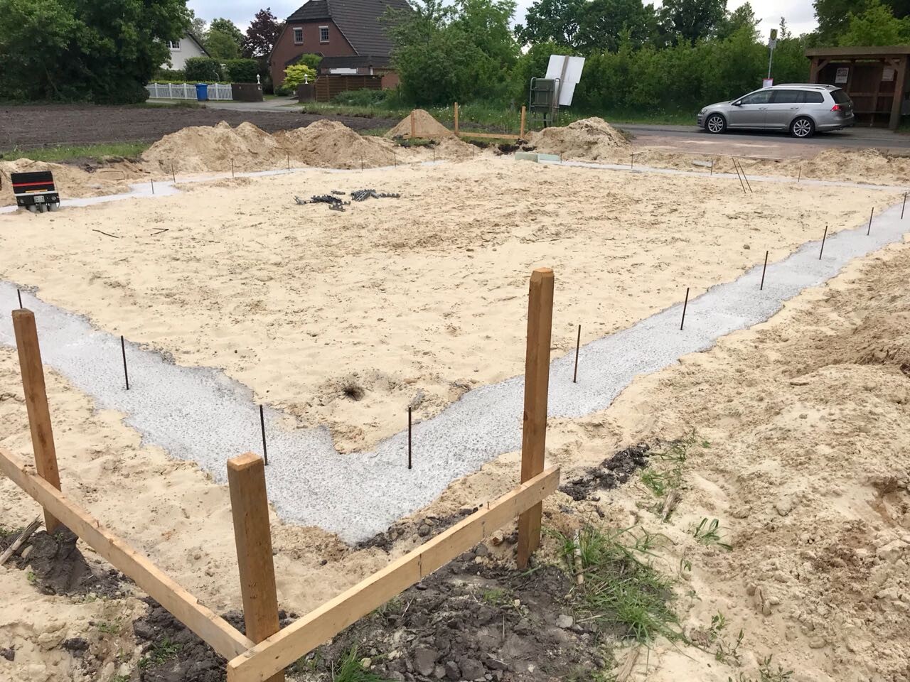 Fundamentbaustelle mit Sand- und Kiesboden, Holzrahmen und Stahlstangen; Auto im Hintergrund.
