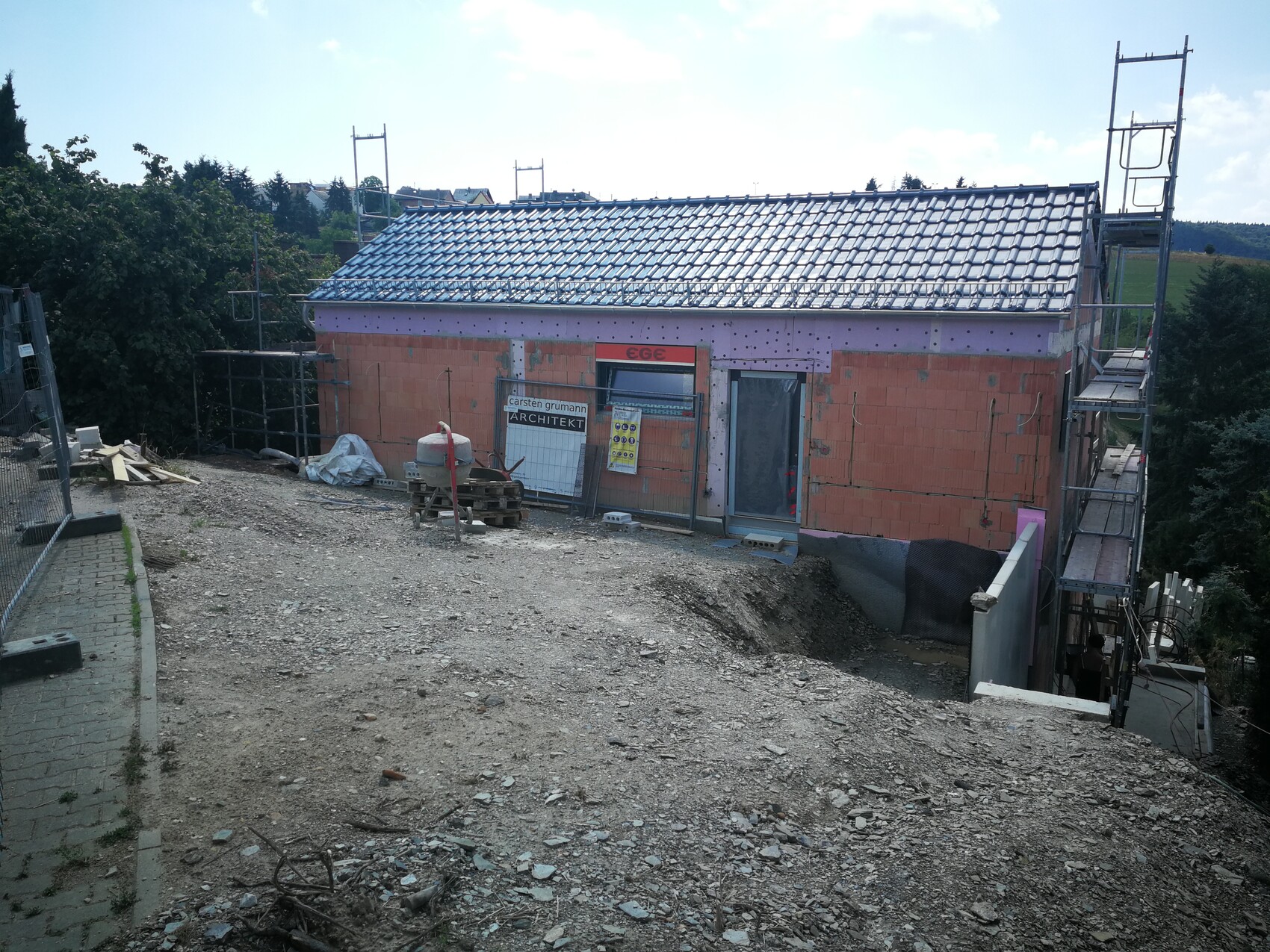Baustellenhaus mit Gerüst, Ziegelwänden, blauem Dach und Baugeräten auf Schotter.