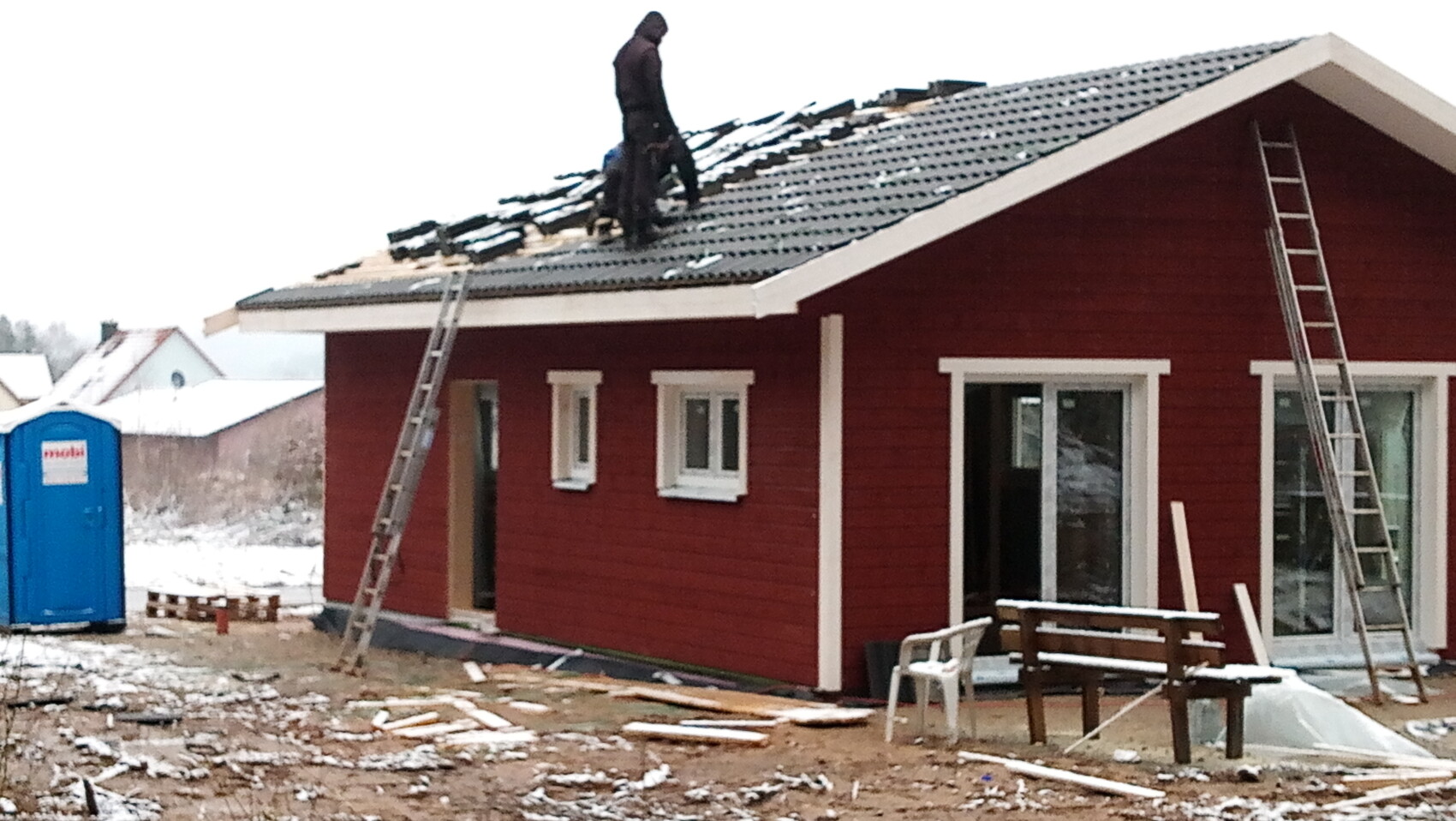 Rotes Holzhaus im Bau; Arbeiter deckt Dach, zwei Leitern, Schnee und Bauholzreste draußen.