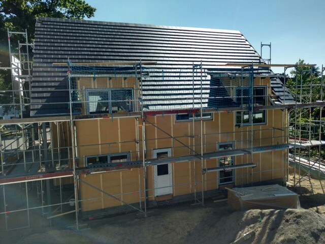 Zweistöckiges Haus im Rohbau, von Gerüsten umgeben, dunkle Dachziegel auf einer Baustelle.