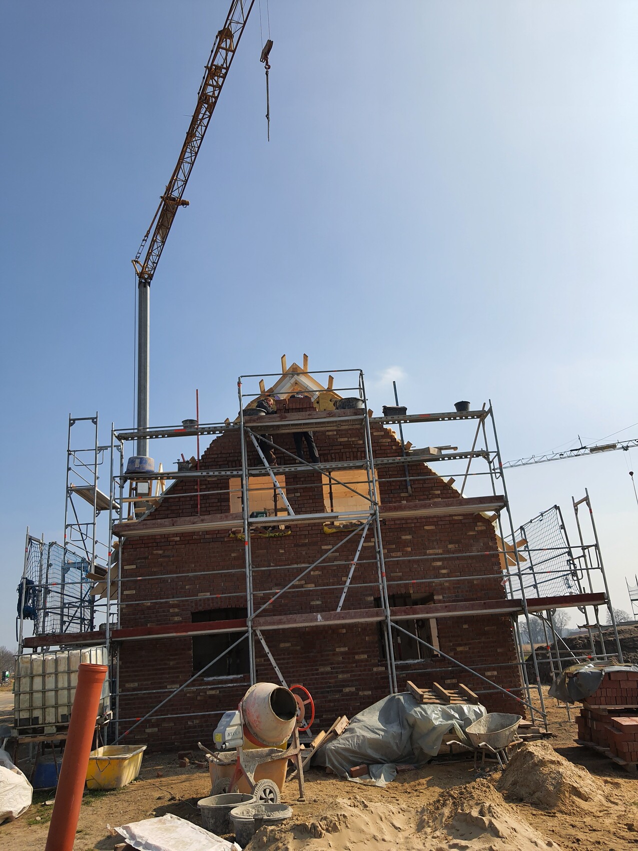 Baukran ragt über Gerüst eines Backsteinhauses; Bauarbeiter arbeiten an der Dachkonstruktion.