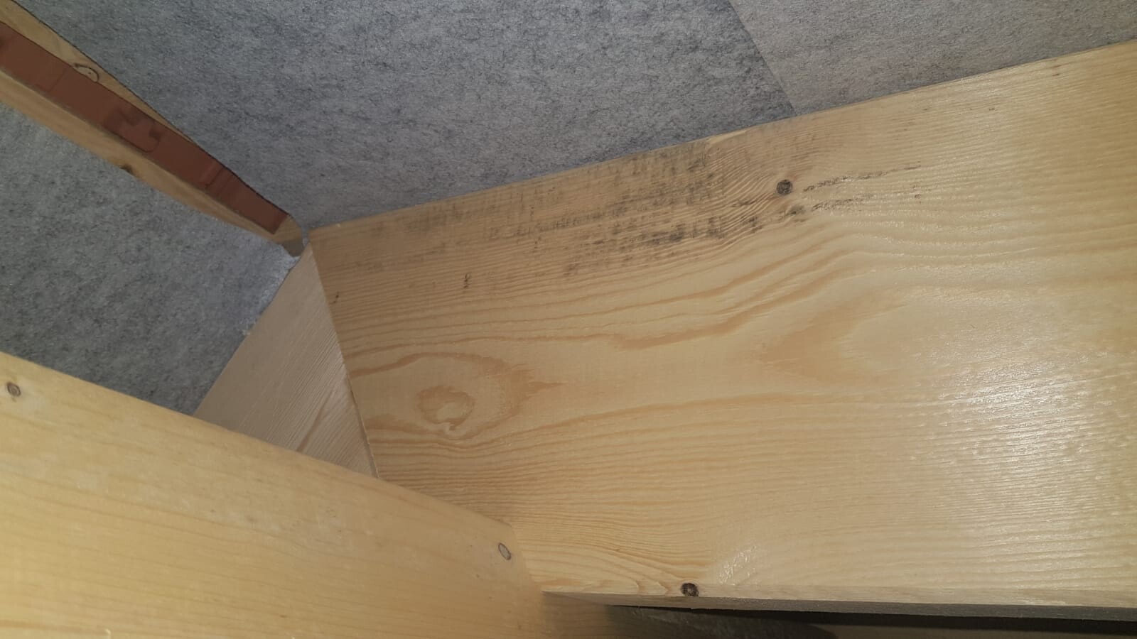 Eckverbindung aus Holzbrettern mit Nägeln; dunkle Verfärbung, grauer Teppich dahinter.