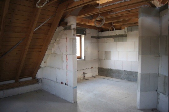 Unfertiger Dachboden mit weißen Blockwänden, Holz-Balken-Dachstuhl und sichtbaren Kabeln.