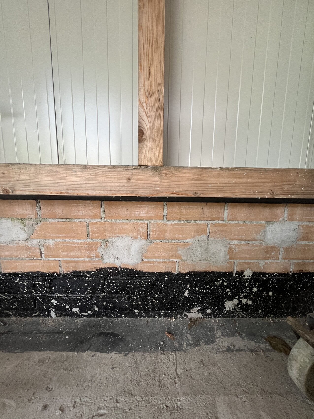 Unterer Wandbereich aus Ziegeln, schwarze Bordüre, Holzpfosten und graue Paneele dahinter.