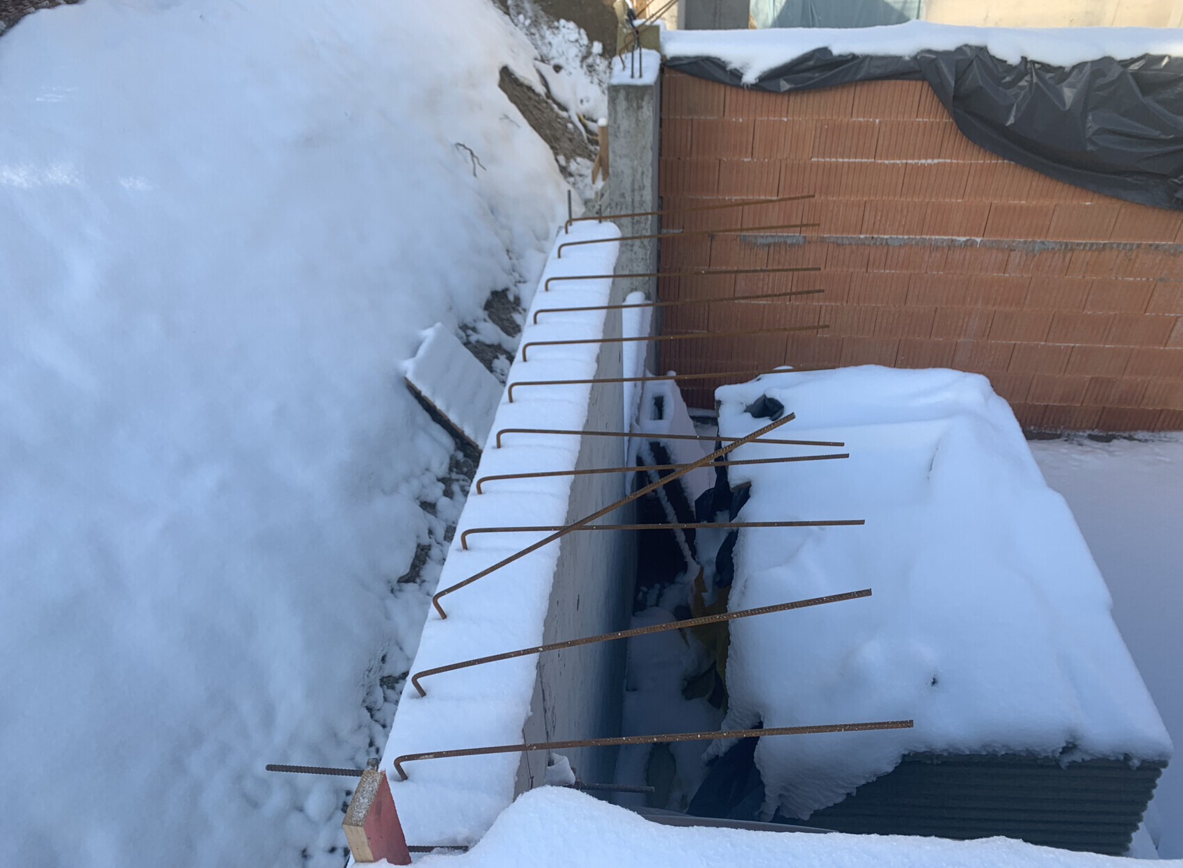 Schnee bedeckte Bauwand mit rostigen Bewehrungsstäben, dahinter eine Ziegelwand.