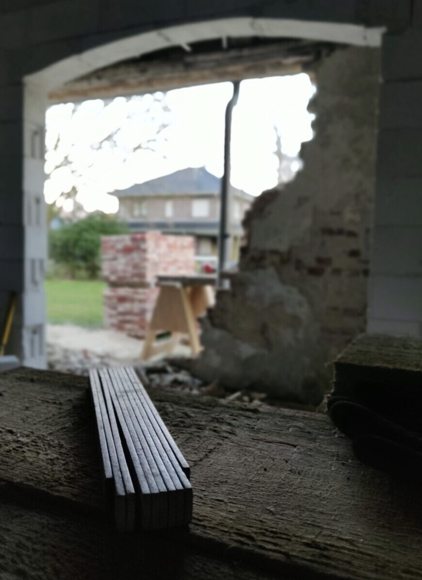 Stapel Metallstangen auf Holzplatte; Baustelle sichtbar durch Wandöffnung, Haus im Hintergrund.