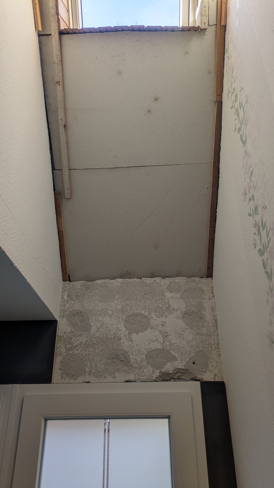 Unfertiger Innenbereich mit rohem Betonboden, offenem Oberlicht, rechte Wand floral tapeziert, Türrahmen unten