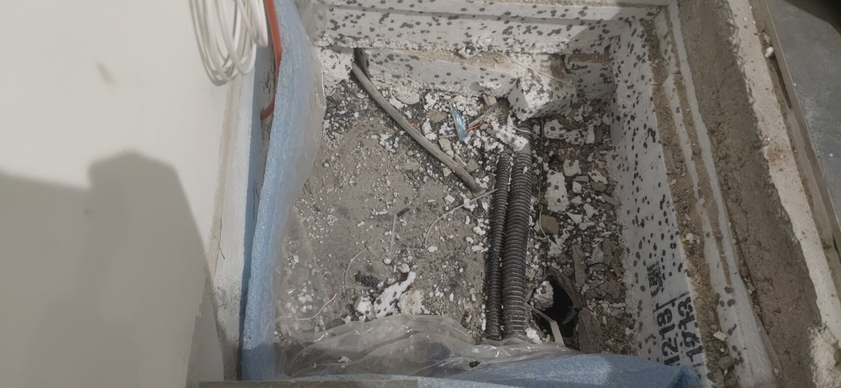 Offene Betonwand mit freiliegenden Kabeln, Rohren und Bauabfällen im Staub.