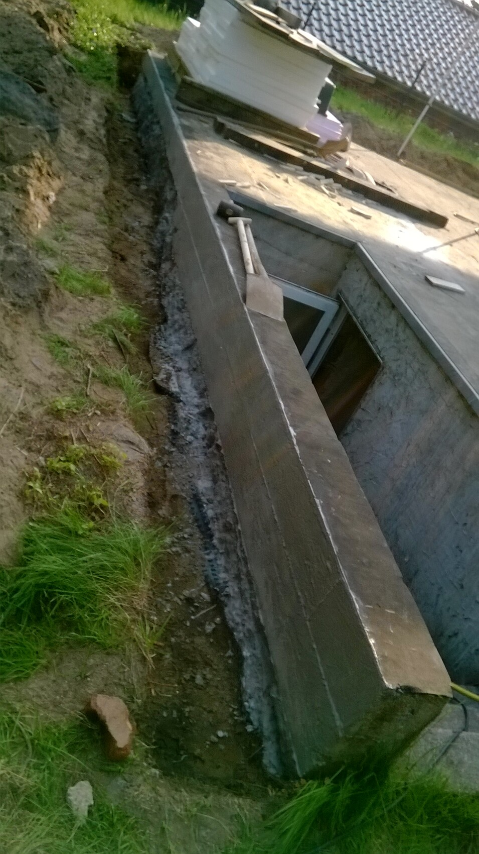 Baugrube mit Betonmauern, offene Kellertüröffnung, Schaufel lehnt an Wand, Bauplatten oben.