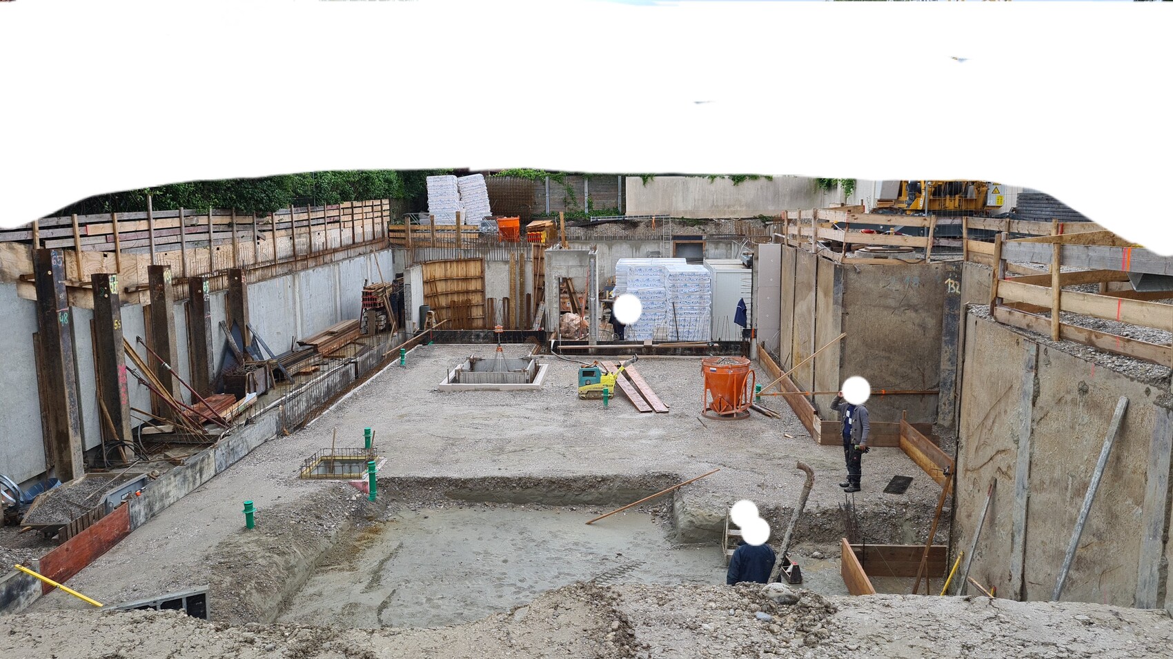 Baustelle: Grube mit Betonwänden, Holzformen, Stahlarmierung; zwei Arbeiter.