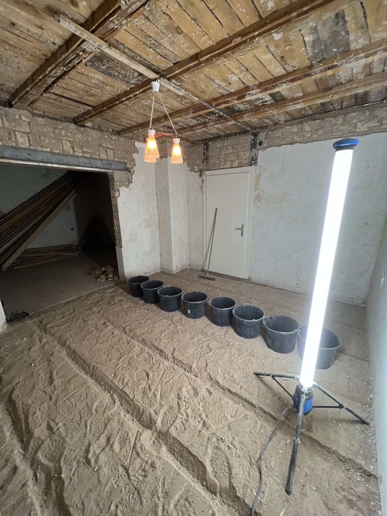 Innenraum einer Baustelle mit Sandboden, Eimern in Reihe, Bauleuchte und Tür.