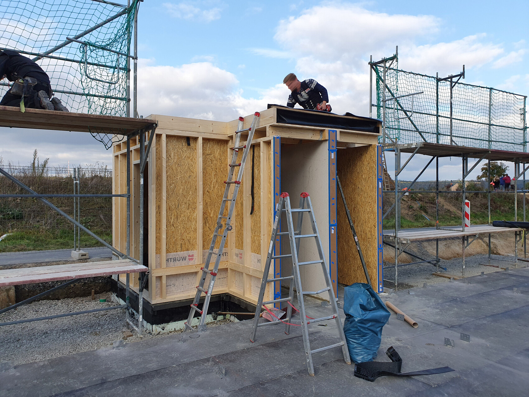 Baustelle: Holzrahmenbau, Arbeiter oben auf dem Dach, Leitern und Gerüst daneben.