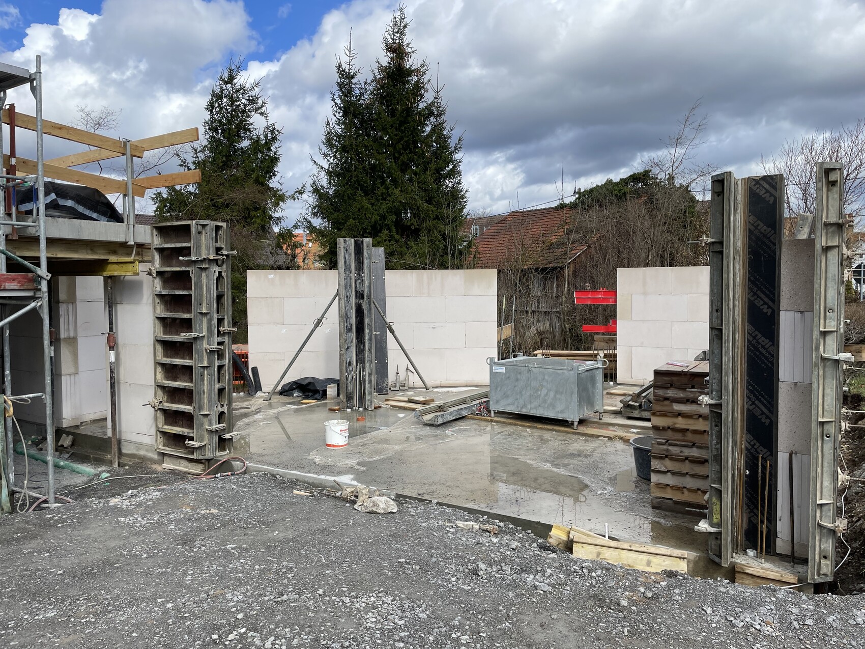 Baustelle mit Betonwänden, Schalung, Stützrahmen, Holzlatten und Baumaterialien sowie Wasserpfützen.