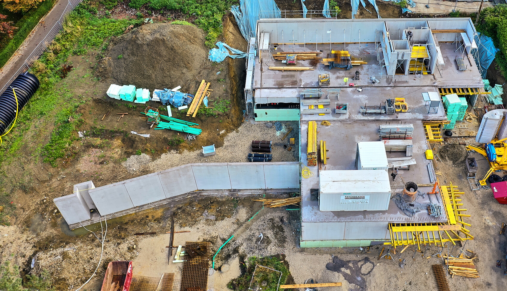 Vogelperspektive einer Baustelle mit unfertigem Gebäude, Materialstapeln, Geländer und Baumaschinen.