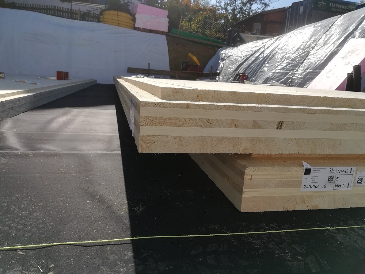 Gestapelte Holzbalken liegen auf einer Fundamentplatte auf einer Baustelle.