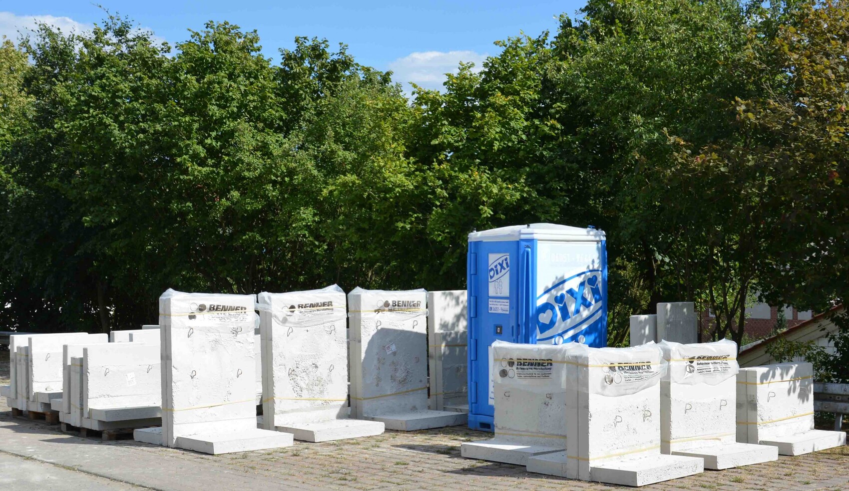 Baustelle mit weißen Steinblöcken, Paletten und blauer Porta-Potty vor Bäumen