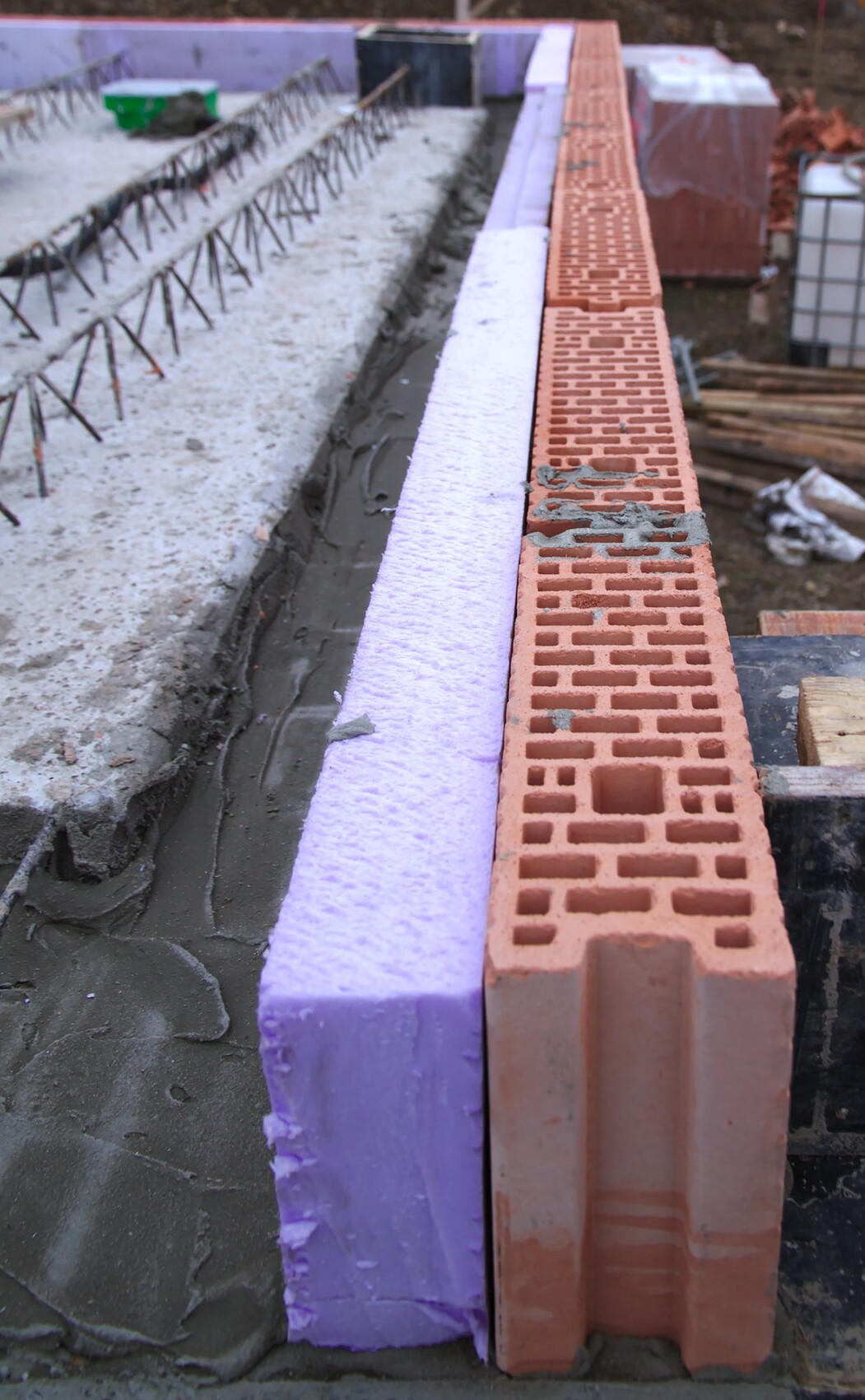 Baustelle mit roten Ziegelsteinen, lila Dämmstoffstreifen, Bewehrungsstahl und feuchtem Beton.