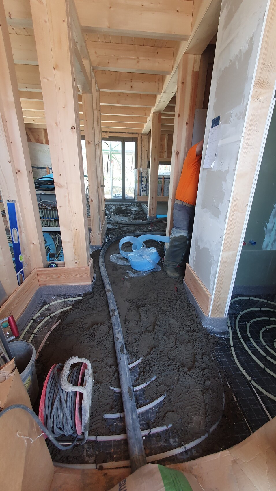 Innenaufnahme einer Baustelle: Holzrahmen, Bauarbeiter in Orange, schlammiger Boden mit Rohren.