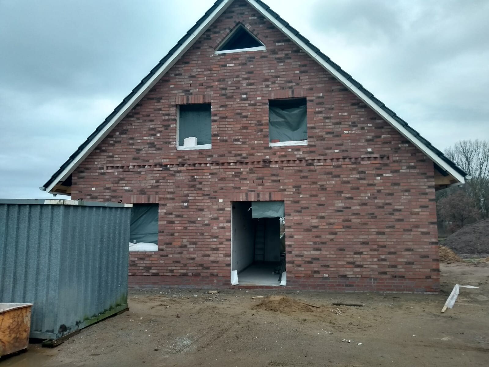 Unvollständiges rotes Backsteinhaus im Bau mit offenen Fenster- und Türöffnungen, Baucontainer links.
