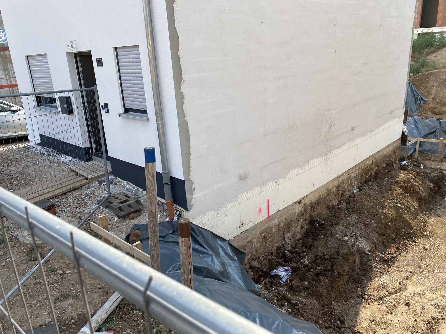 Baustelle neben weißem Haus: Graben am Fundament, Bauzaun im Vordergrund.