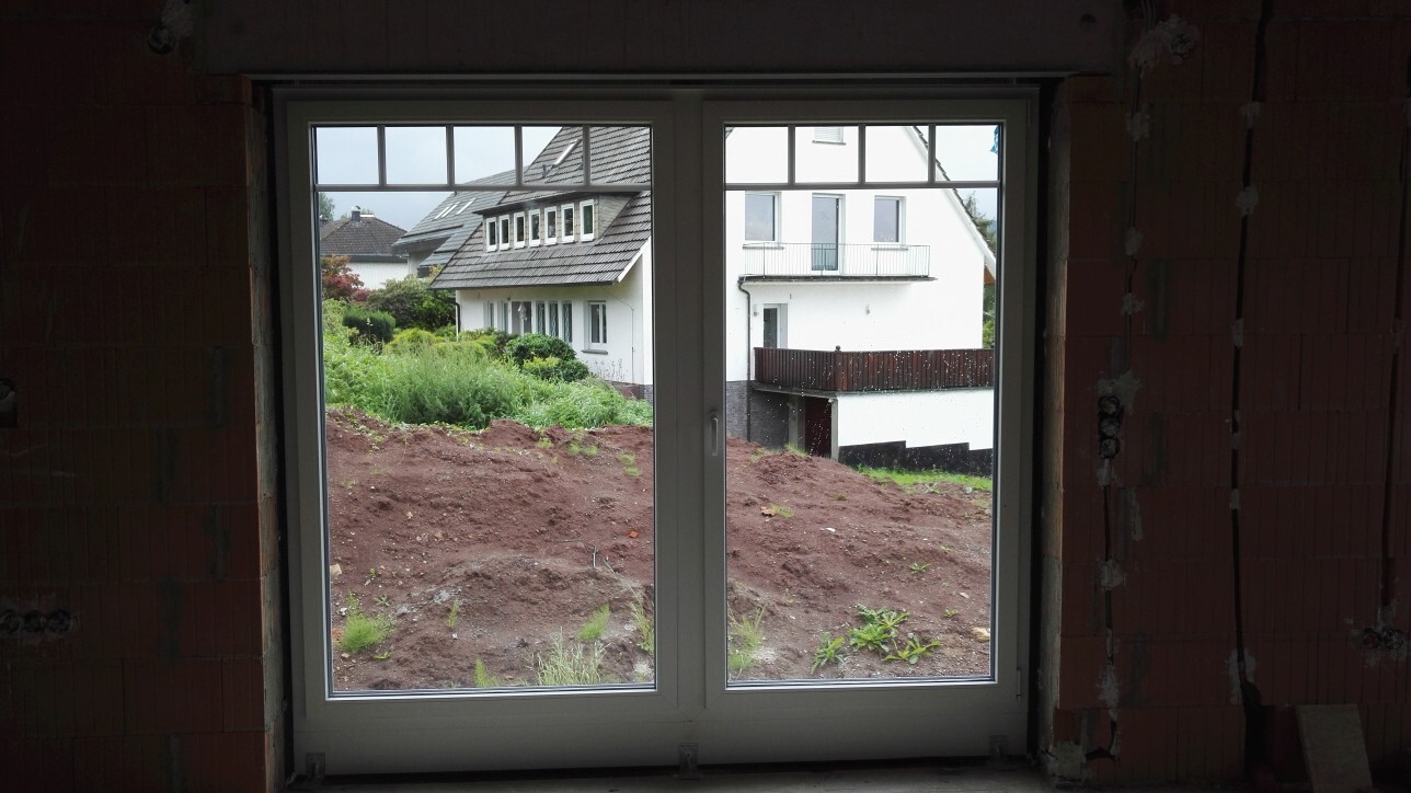 Blick durch großes Fenster auf Baustelle: roher Innenbereich und fertiges Haus im Hintergrund.