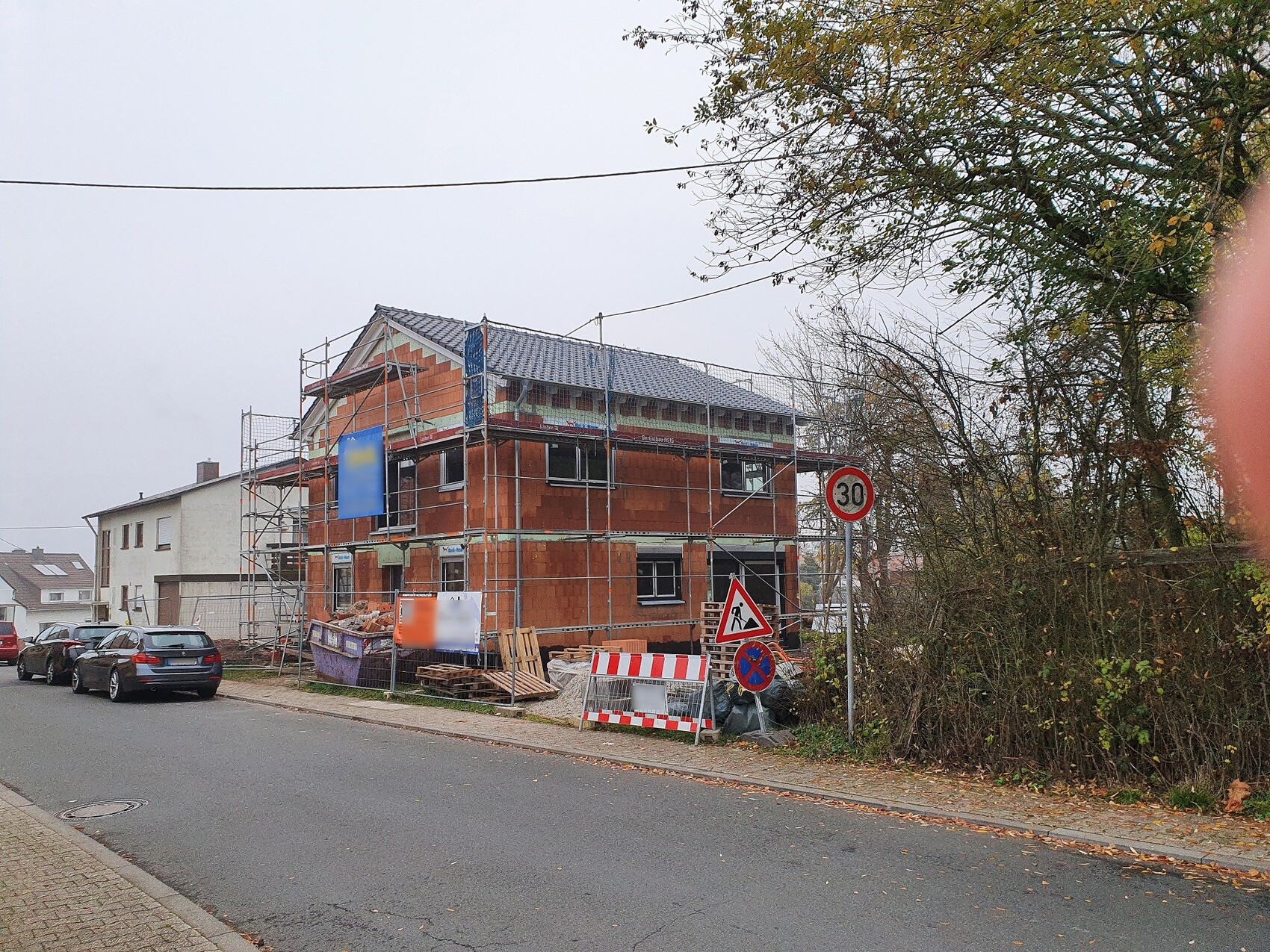 Rohbau eines zweistöckigen Hauses mit Gerüst an der Straße, Bauholz, Schilder, parkende Autos.