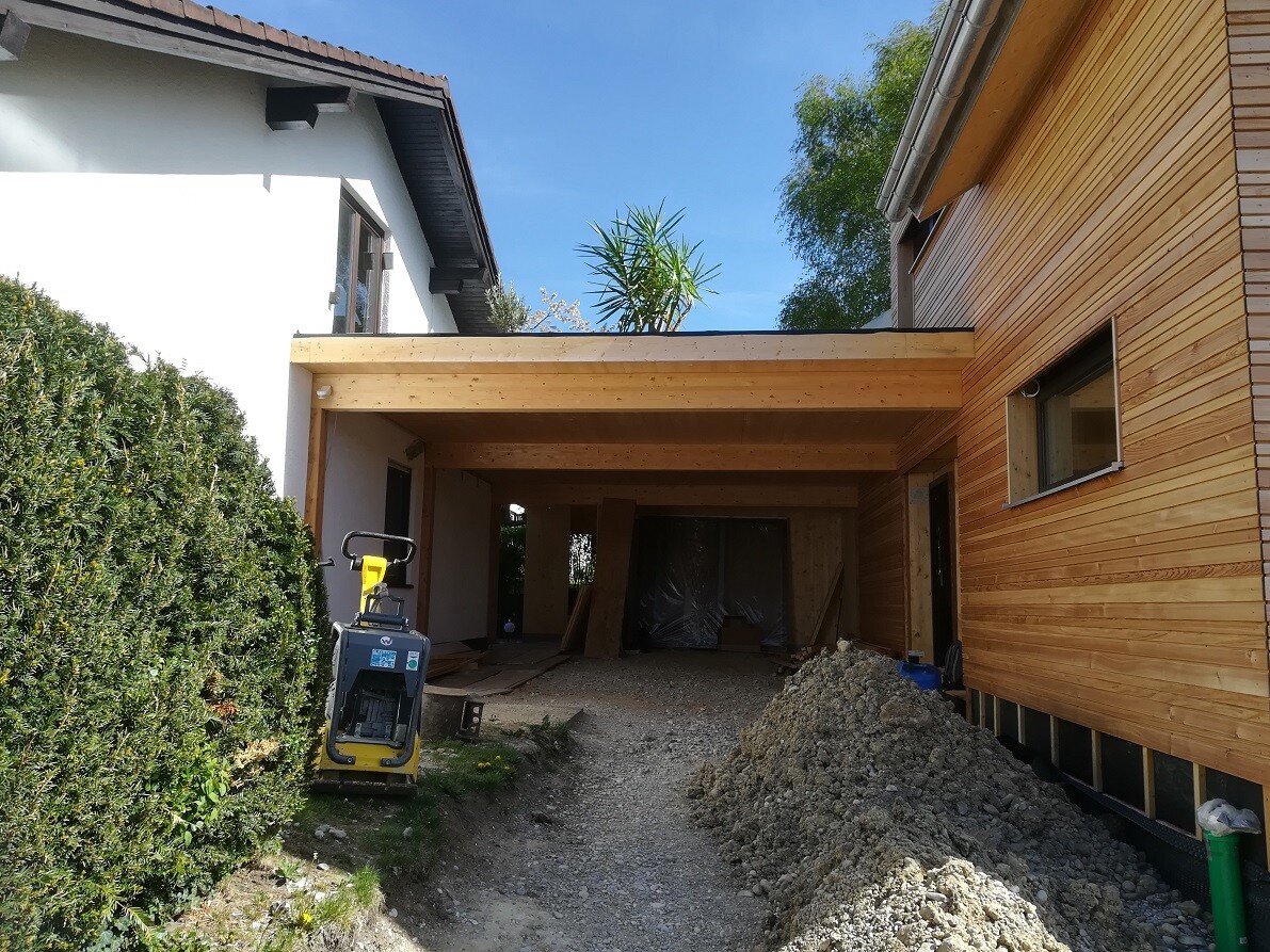 Baustelle mit holzverkleideten Anbau, Kieshaufen, Baugerät, grüne Hecke links, blauer Himmel.